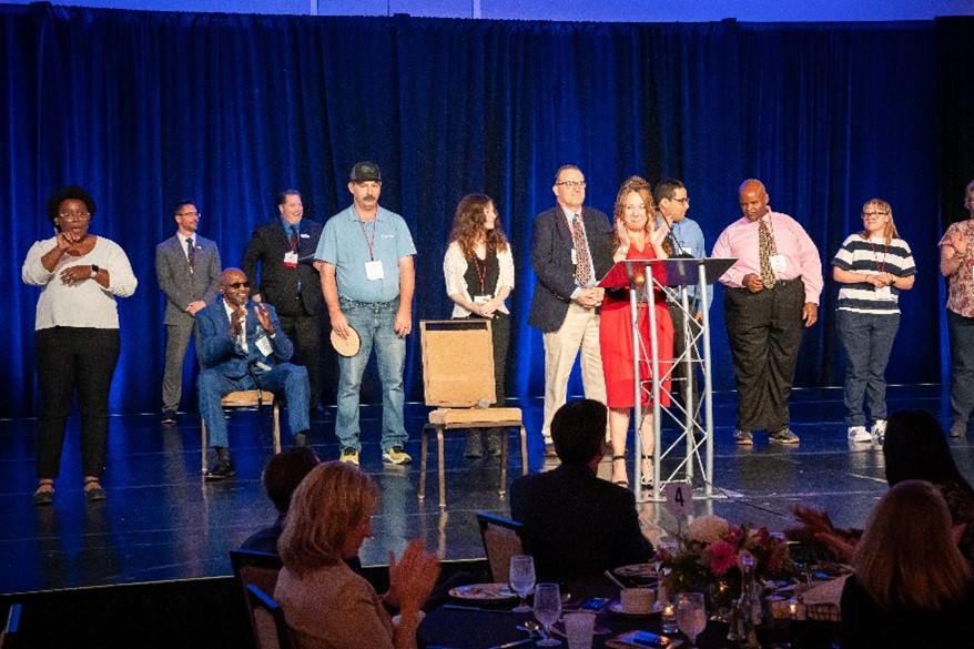 A group of the 2024 Achievement Award winners are standing on a large stage as they receive their award.